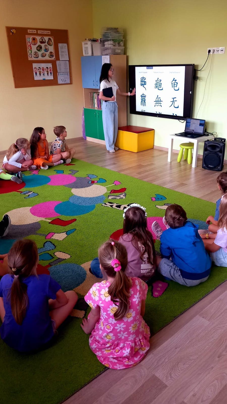 An exchange participant teaching a group of children sitting on a classroom carpet, with characters on the screen